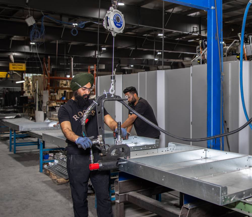 Malach Metal’s pool panel manufacturing line, with stud welding, clinching, riveting, and fastening capabilities Two men wearing blue overalls operate a machine, focused on their task in an industrial environment.